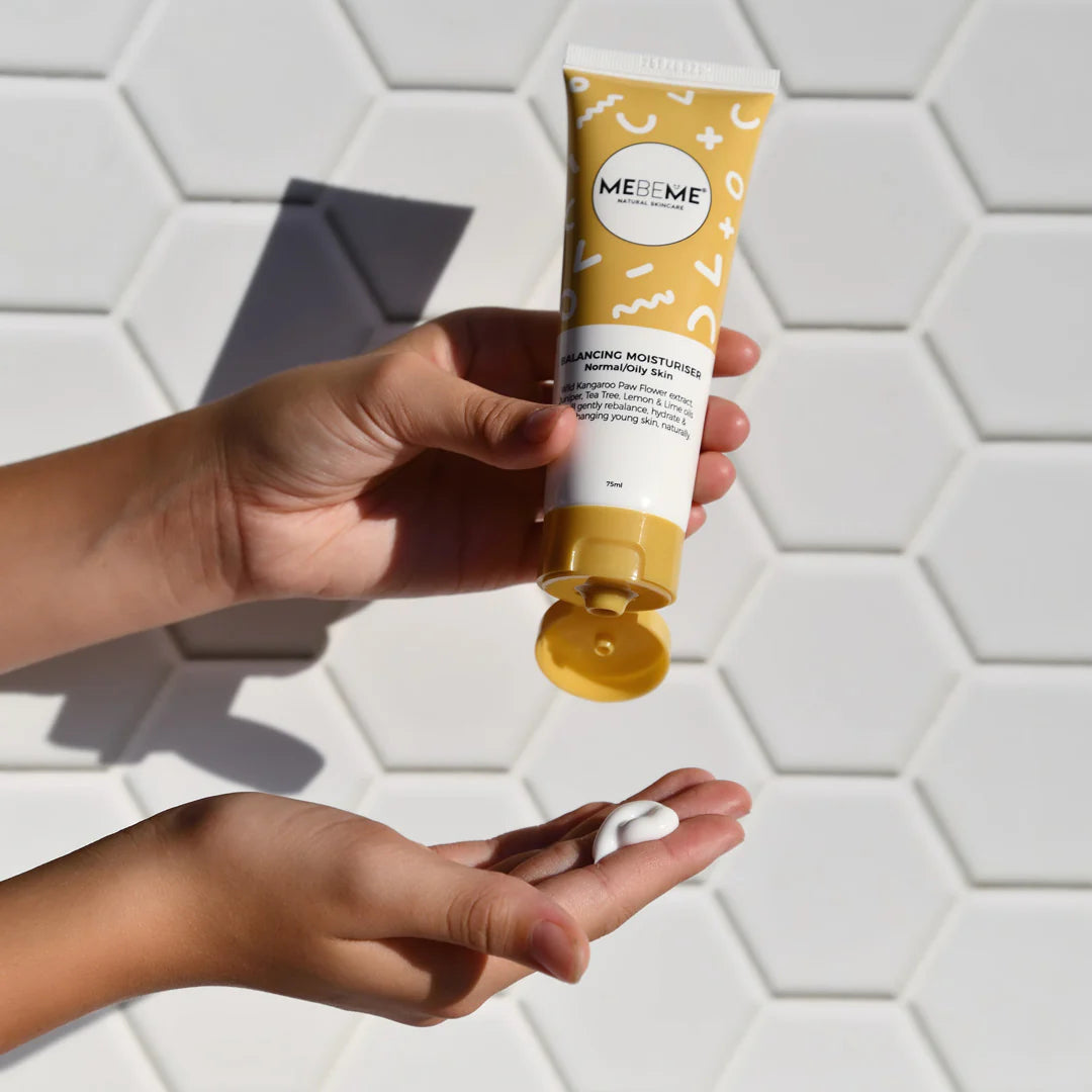Hand holding a tube of MEBEME moisturiser against a white tiled wall.