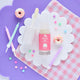 A photo of a birthday cake-scented natural lip gloss for kids, displayed on a white flower shape with pink chequered material and purple background. The lip gloss is presented in a clear tube with a white cap, and the packaging is also shown with birthday themed decorations.