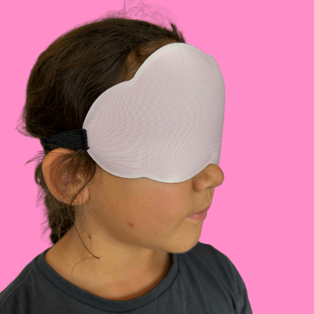 The profile of a little girl wearing a cloud shaped sleep mask against a pink background