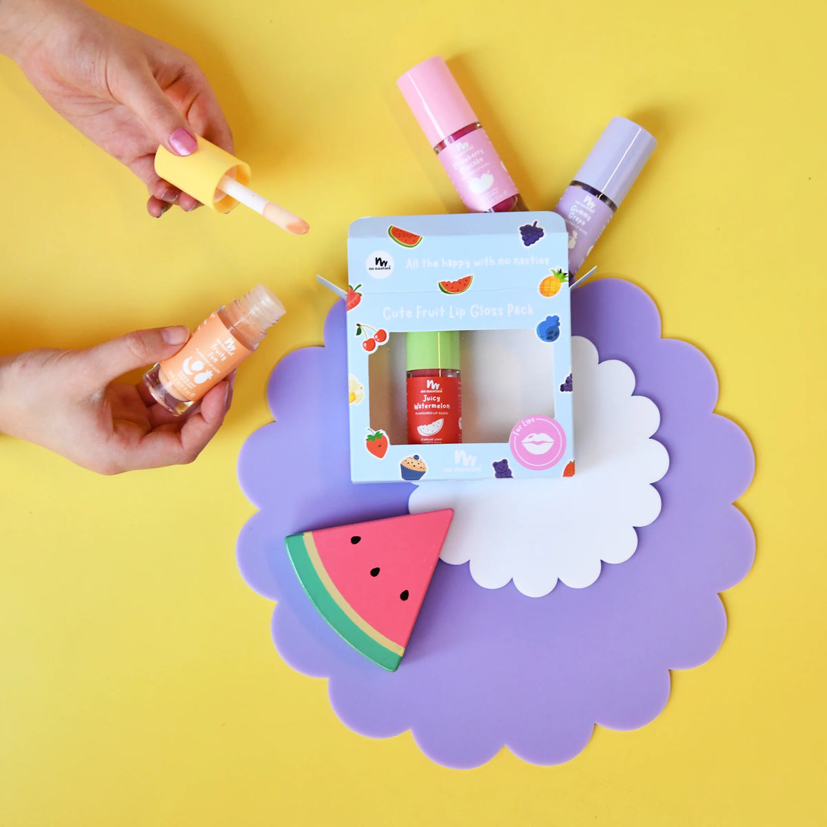 A beautiful gift set featuring a 'Cute Fruity Lip Gloss Pack' displayed on a purple scalloped card over a yellow background. Hands are shown holding two of the lip products (one with a yellow applicator, one with an orange applicator) alongside the boxed set and a watermelon shaped item.