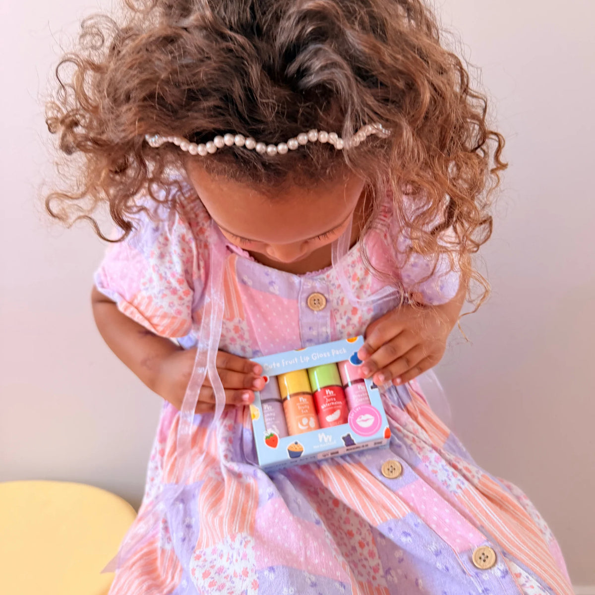 A toddler girl with curly brown hair wearing a pearl headband and a pink patterned dress, looking down at a small box of colorful lip balm tubes held in her hands.