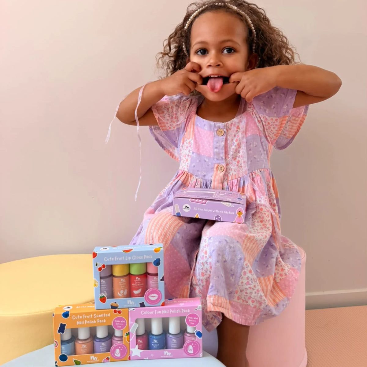 A young child wearing a pastel patchwork dress sits on a pink stool, playfully sticking their tongue out. In front of them are three colorful boxes of nail polish sets