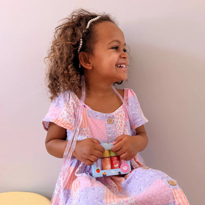 A young girl with curly brown hair wearing a lavender and pink patterned dress and a pearl headband, smiling while holding a small set of lip balm tubes against a plain light wall.