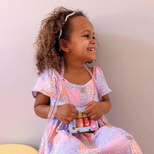 A boxed lip gloss pack surrounded by colorful fruit stickers on a lavender background. The package reads 'Cute Fruit Lip Gloss Pack' and contains four gloss bottles with fruit themed labels: juicy watermelon, strawberries cupcakes, fruity fun, and cucumber grape.