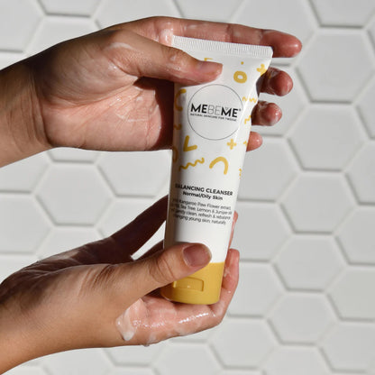 Hand holding a tube of MEBEME cleanser with soap suds on a white background