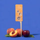 A product shot of a biodegradable styling comb on a blue background, styled with peaches and a plum. The comb features a bright orange card insert with white text and illustrations, and a light beige plastic comb head on a white stick.