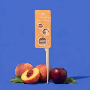 A product shot of a biodegradable styling comb on a blue background, styled with peaches and a plum. The comb features a bright orange card insert with white text and illustrations, and a light beige plastic comb head on a white stick.