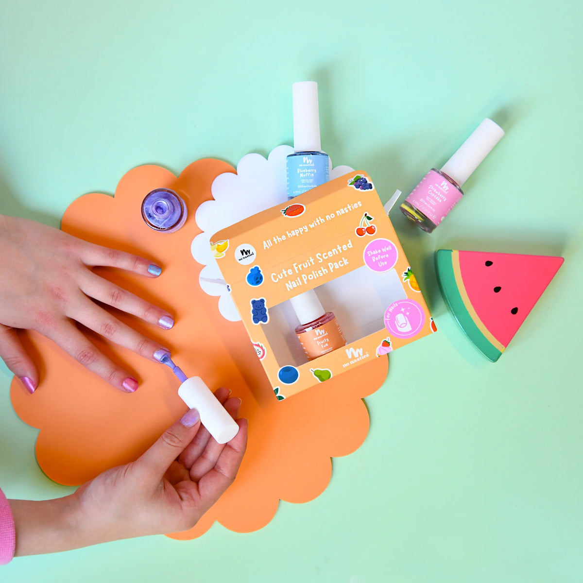 A colorful nail polish set displayed on a mint green background, featuring a hand with lavender painted nails near an orange scalloped mat. The set includes a box labeled 'Cute Fruit Scented Nail Polish Pack', several nail polishes in blue, pink, and orange, and a watermelon shaped item.