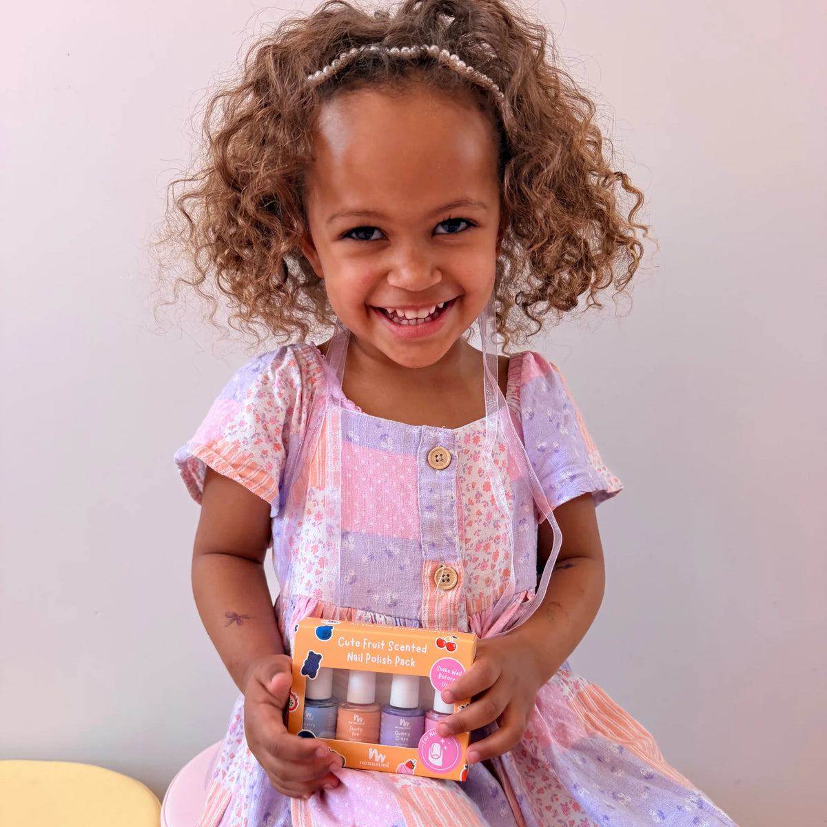 A young girl with curly brown hair wearing a pink and purple patchwork dress holds a box of nail polish in a warm, indoor setting.