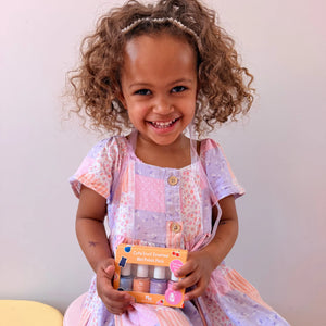 A young girl with curly brown hair wearing a pink and purple patchwork dress holds a box of nail polish in a warm, indoor setting.